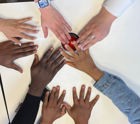 8 Hände von Menschen unterschiedlicher Hautfarbe, versuchen einen roten Buzzer in der Mitte eines Tischs zu drücken.